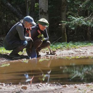 Two women crouching near a large puddle in a forest. 
