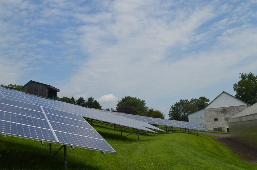 Several solar panels line a grass field with a house in the distance.