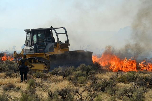 A person dressed in protective fire gear walks toward a bulldozer parked in front of a wildfire.