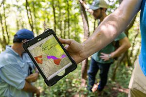 A man holds out a tablet showing his location and the high elevation of the forest that a small group of people stands in.