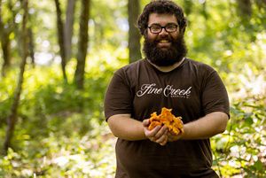 A smiling man poses in a shaded forest clearing holding a handful of bright orange chanterelle mushrooms.