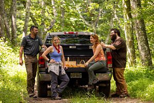 Four people sit and stand together around the bed of a pickup truck parked on a wide forest trail.