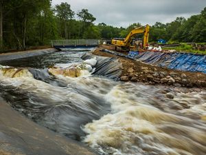 Water rushes down a channel in the foreground, while a large, yellow machine works to demolish a dam in the background.