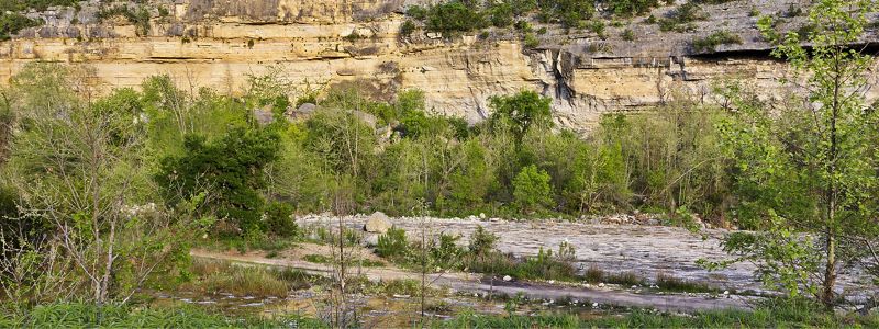 Water runs along the base of a limestone cliff covered in trees.