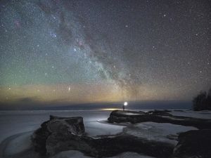A person holds a light as they stand on a rock formation. The sky if filled with millions of stars and the hues vary.