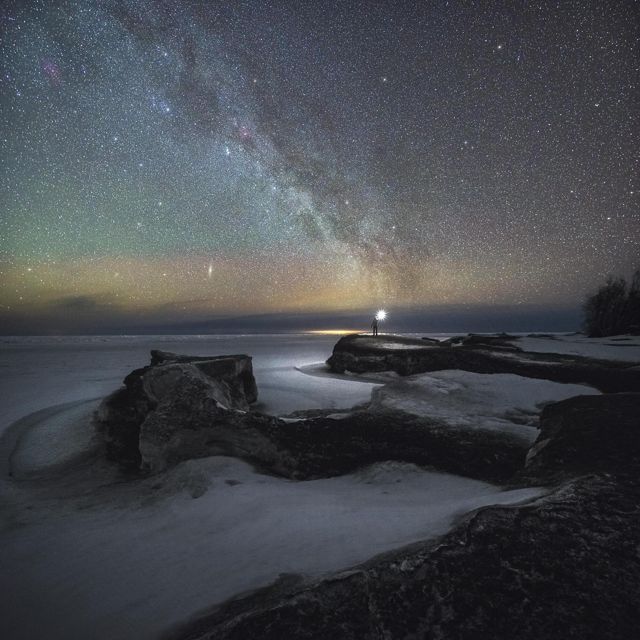 A person stands on the edge of the frozen water and holds a light. The sky is lit up with stars. 