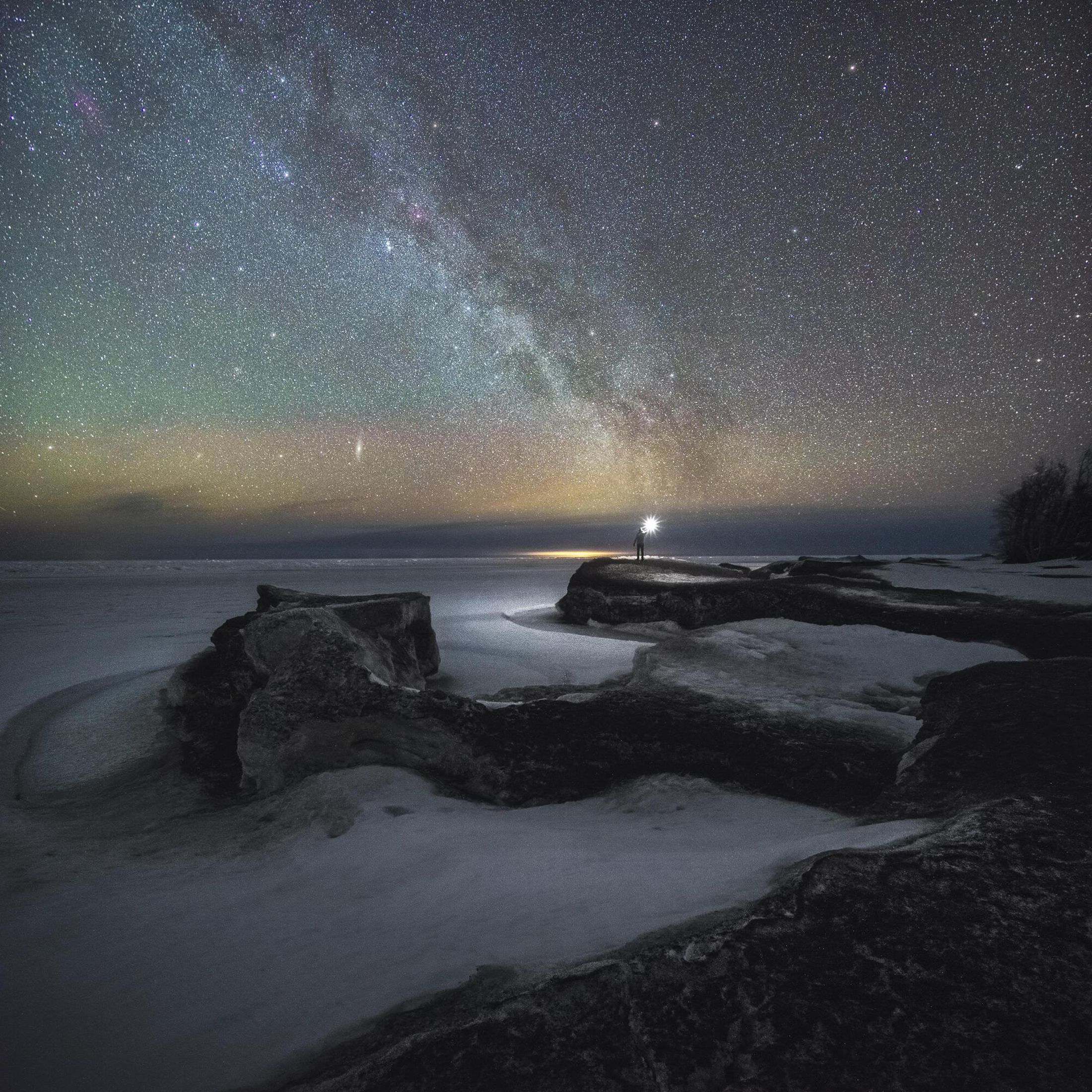 A person holds a light as they stand on a rock formation. The sky if filled with millions of stars and the hues vary. 