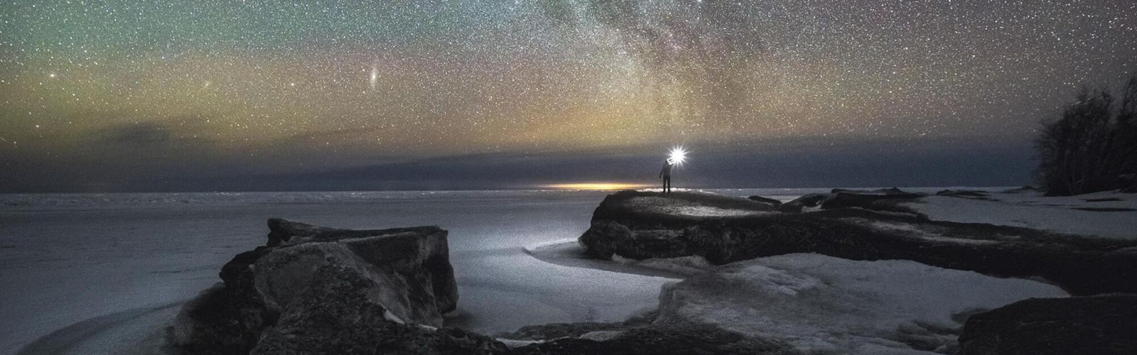 A person holds a light while standing at the edge of a lake. The sky is covered in thousands of stars. 