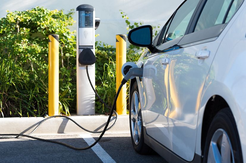 An electric car is parked at a charging station. A large plug is inserted into the front quarterpanel of the vehicle.