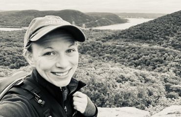 A person take a selfie in front of a vista view of a winding river curving around the base of a small mountain.
