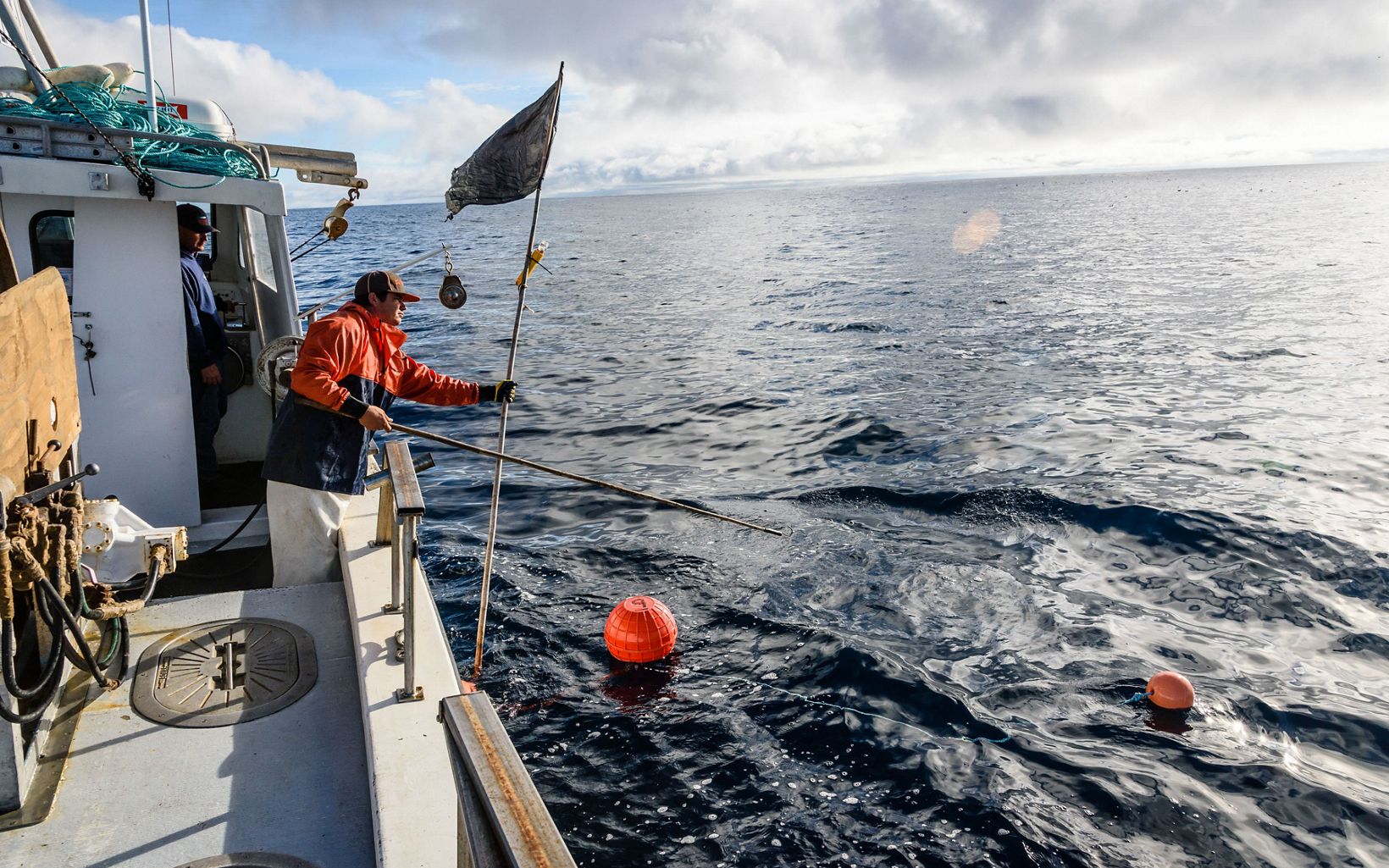 Fisherman holding a flag and stick, leaning over the side of a boat with orange buoys floating the water.