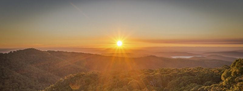 A couple sits on a rock and watches as sun rises over forested hills in the distance.