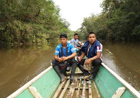 3 people sit on one end of a boat on the river.
