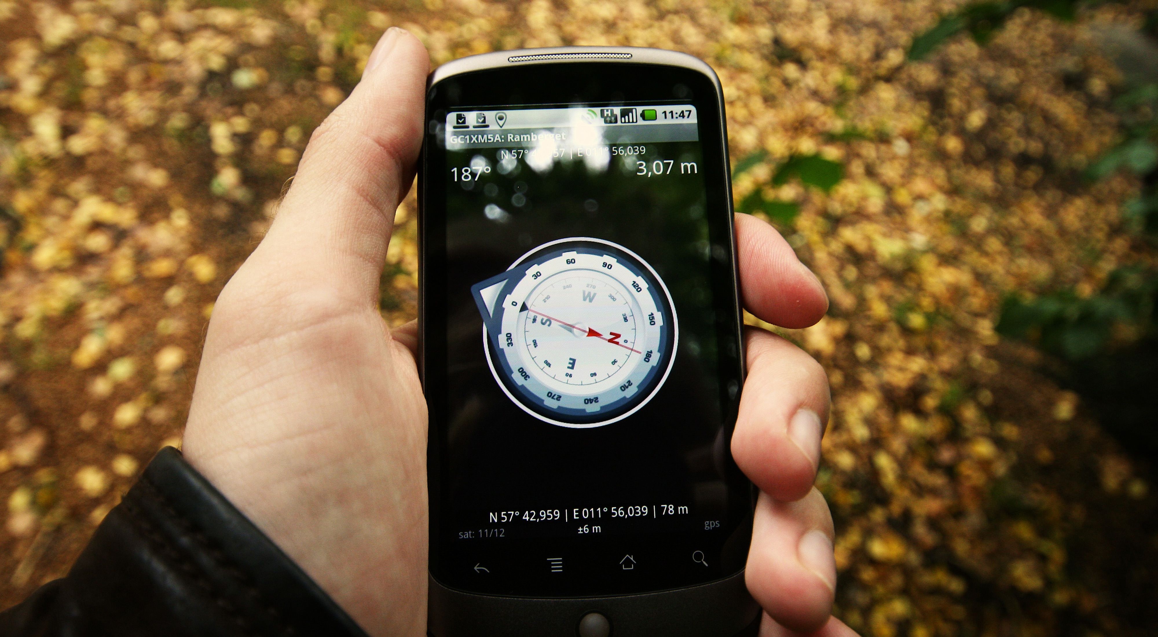 A man standing outdoors holds a cell phone showing map coordinates on its screen.