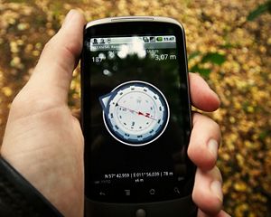 A man standing outdoors holds a cell phone showing map coordinates on its screen.