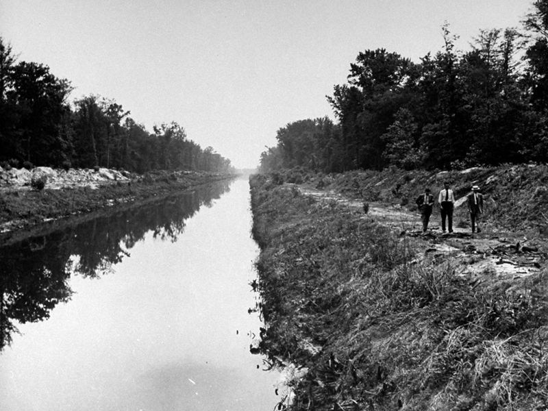 Black and white photo taken in 1943. Three men walk along a narrow river channel. The banks on either side are piled with soil forming high berms.