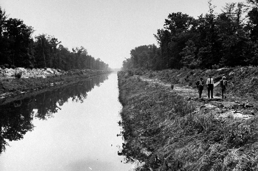 Black and white photo taken in 1943. Three men walk along a narrow river channel. The banks on either side are piled with soil forming high berms.