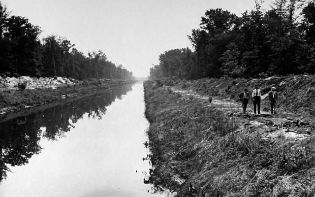 Black and white photo taken in 1943. Three men walk along a narrow river channel. The banks on either side are piled with soil forming high berms.