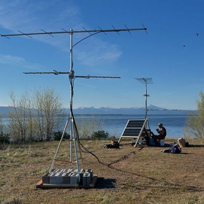 A piece of equipment with poles stands in a grassy area, while a person sits in the distance with a solar panel, with a mountain and water in the background.