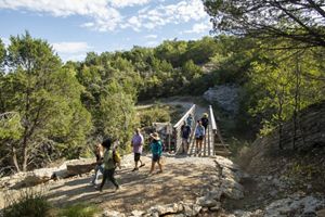 A group of men and women hike along a bridge leading to a rocky trail surrounded by dense green brush.