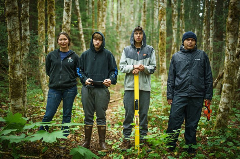 4 jóvenes adultos con botas y chaquetas se encuentran en un bosque con diversas herramientas.