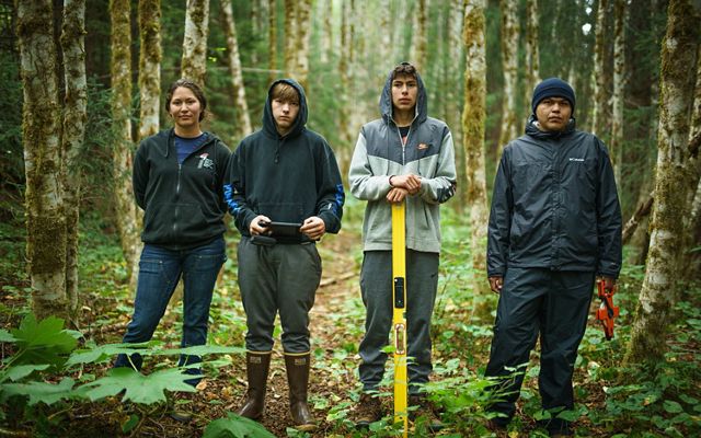 4 jóvenes adultos con botas y chaquetas se encuentran en un bosque con diversas herramientas.