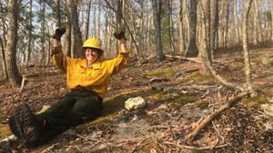 A woman wearing yellow fire gear sits on the ground in a forest. She is smiling and her arms are raised over her head in an expression of celebration.
