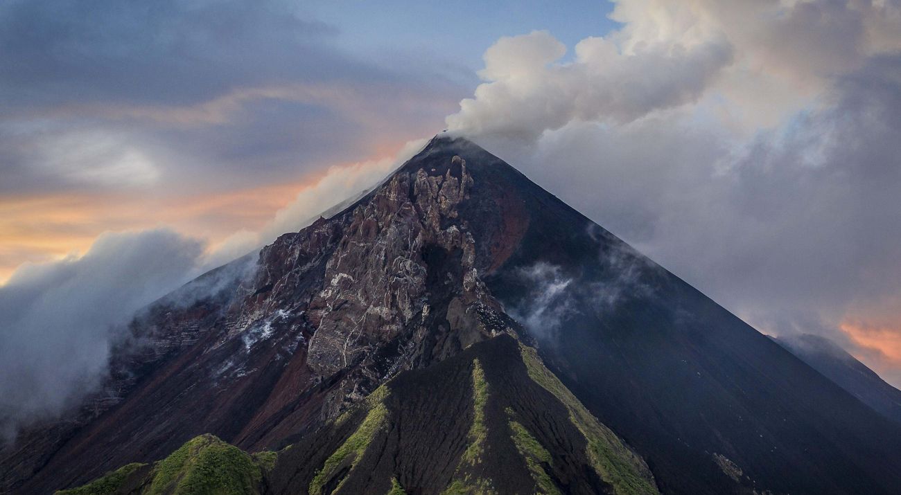 White clouds rise from the peak of a mountain at sunset.