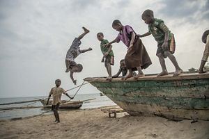 Several small children stand on the edge of a wooden boat on a beach. One of the children has jumped off the boat and is upside-down in midair.