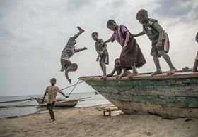 Several small children stand on the edge of a wooden boat on a beach. One of the children has jumped off the boat and is upside-down in midair.