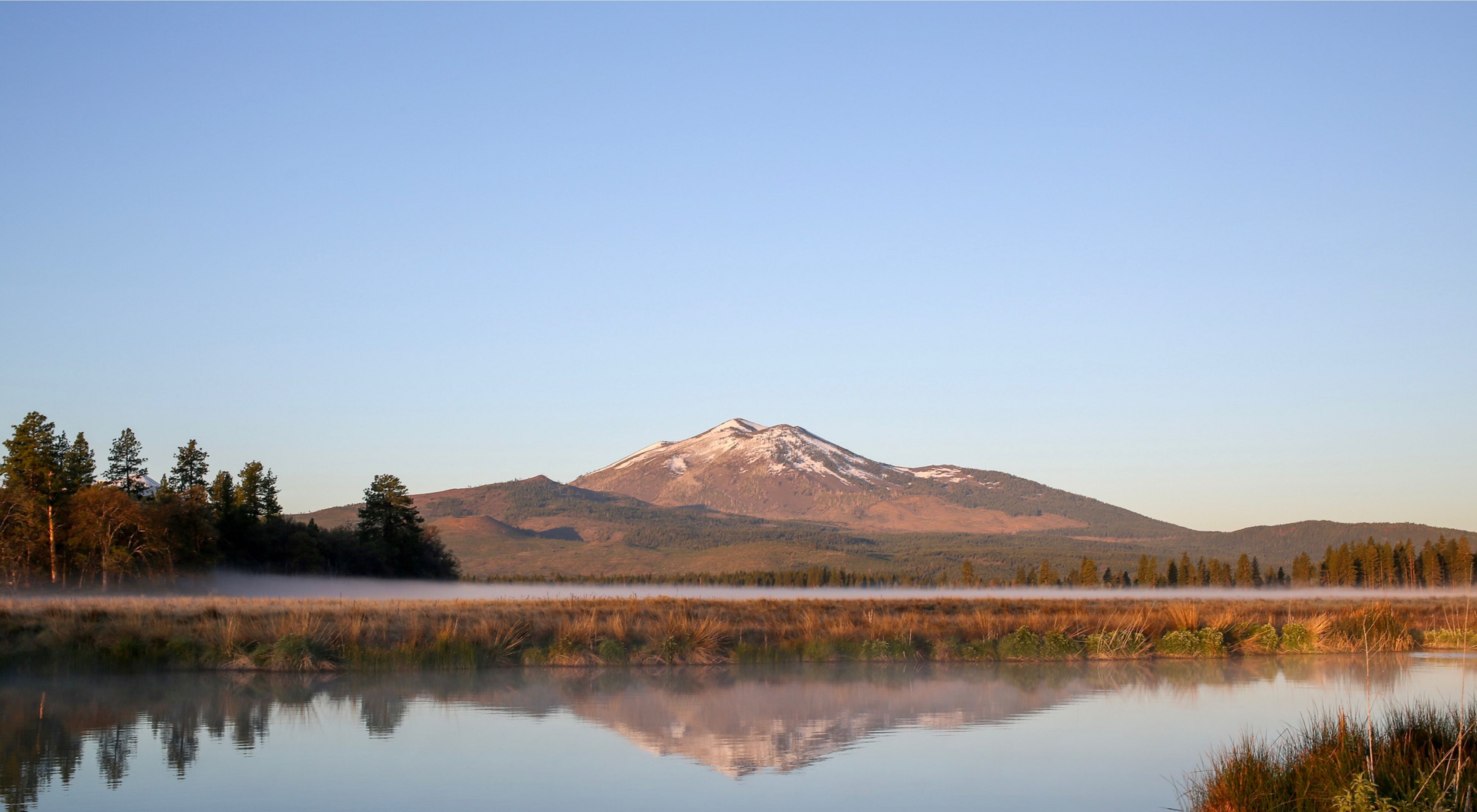 Glassy water with grasses on both sides in the foreground and a snowy mountain in the background. 