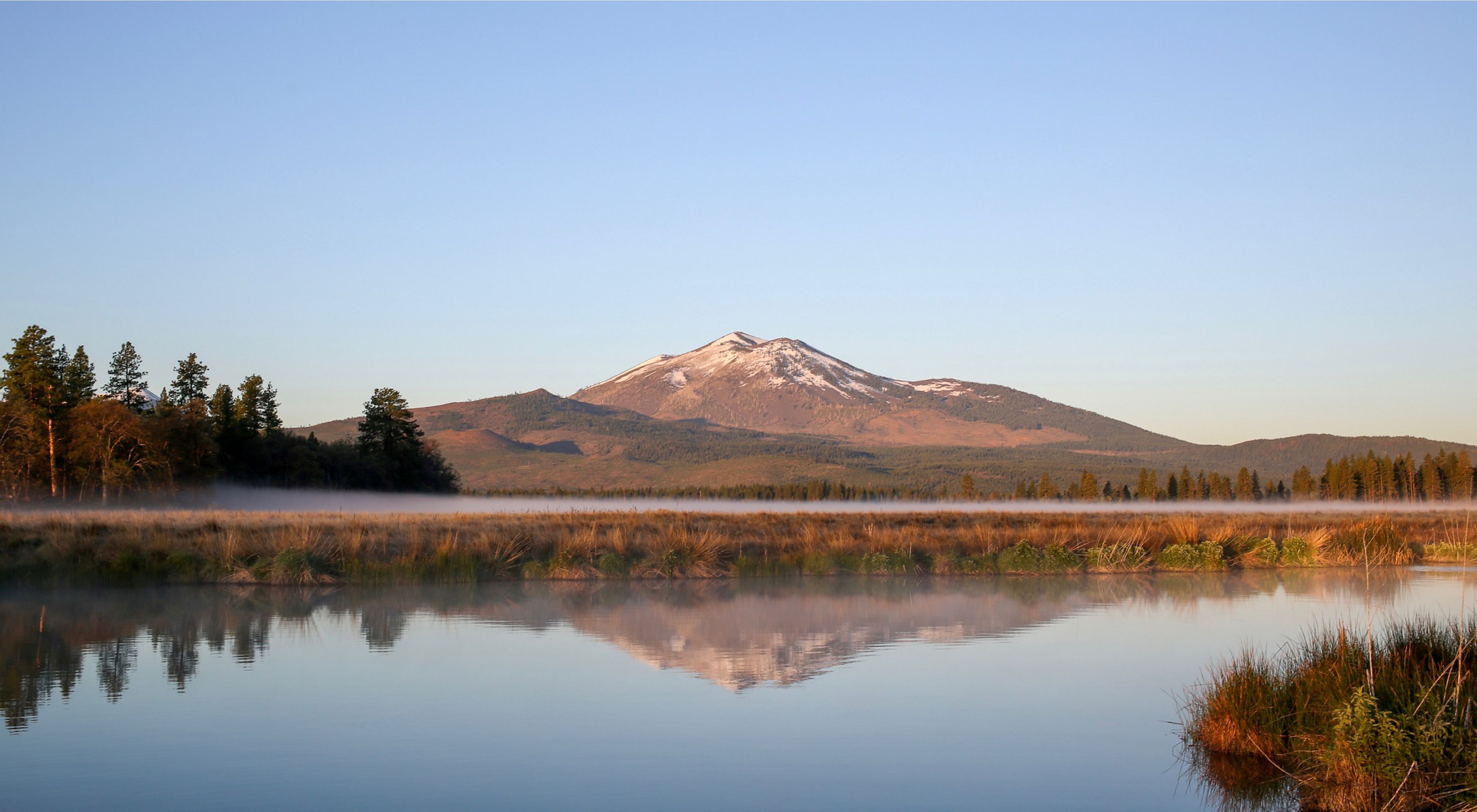 Glassy water with grasses on both sides in the foreground and a snowy mountain in the background. 