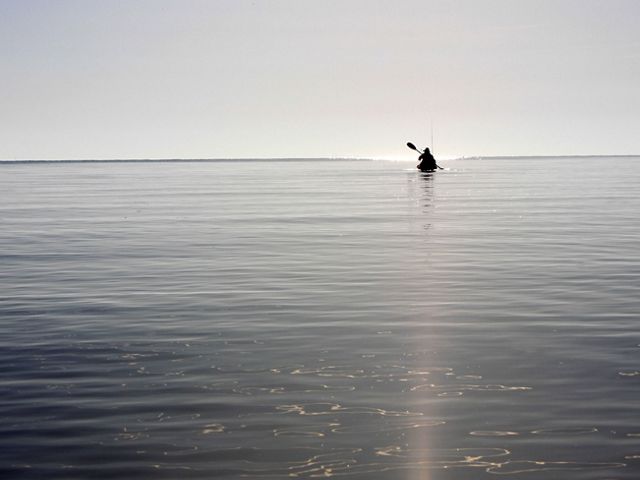 A person floats in a kayak in a flat, wide body of water. They are silhouetted against the sky and seem to hover in the distance at the horizon.
