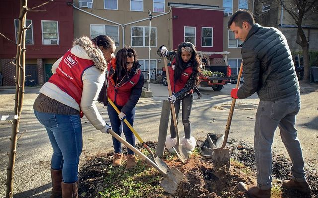 Four people in vests hold shovels and dig into a dirt patch in a paved lot.