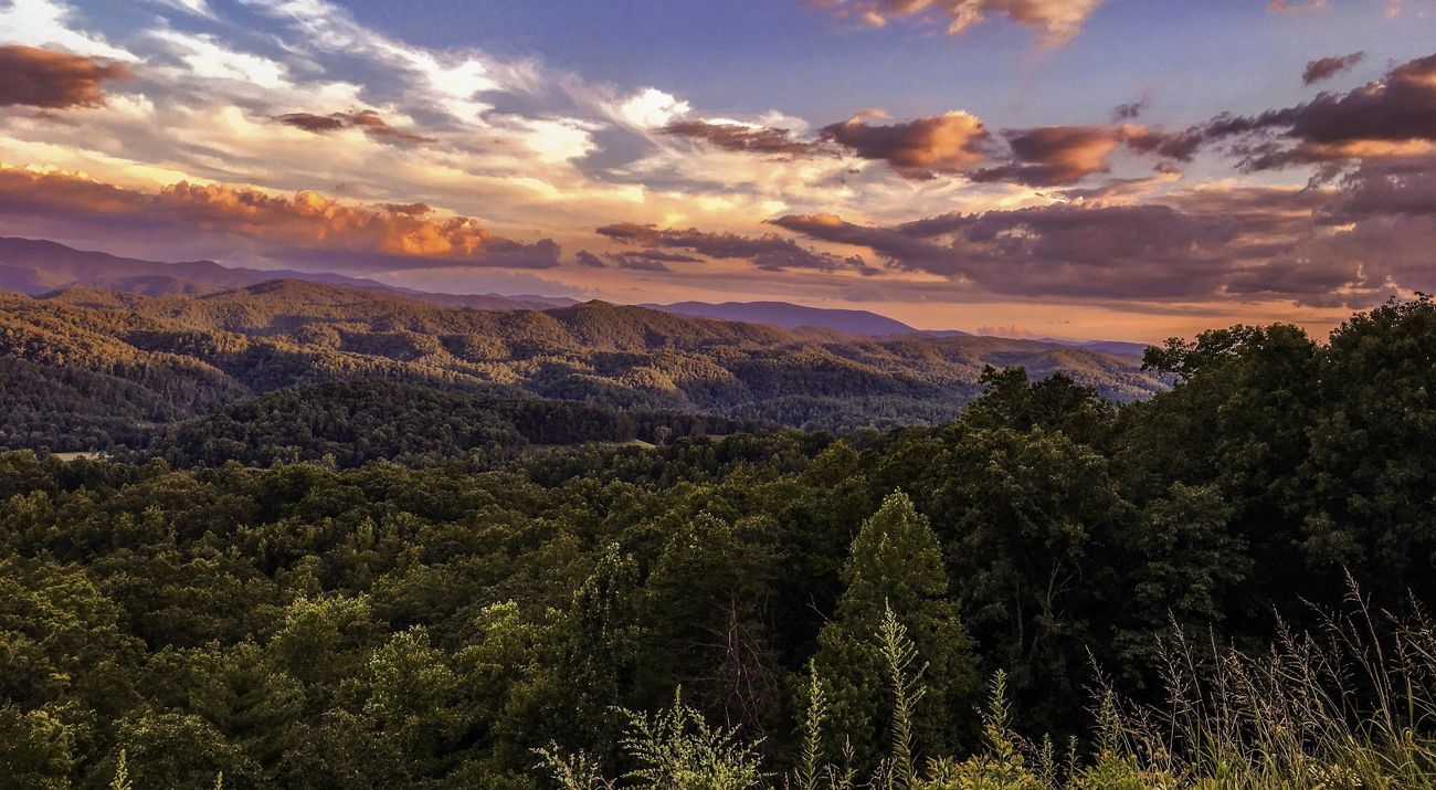 Rolling mountains covered in different shades of green trees stand in the foreground of a cloudy blue sky.