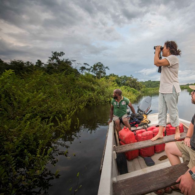 Three people on a boat on a body of water, one stands with binoculars looking out into a lush vegetated area.