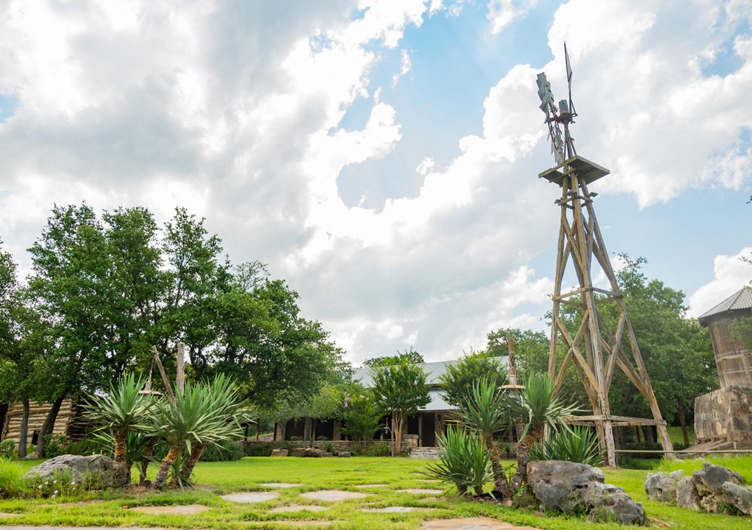 A windmill looms in front of a house surrounded by green grass and trees.