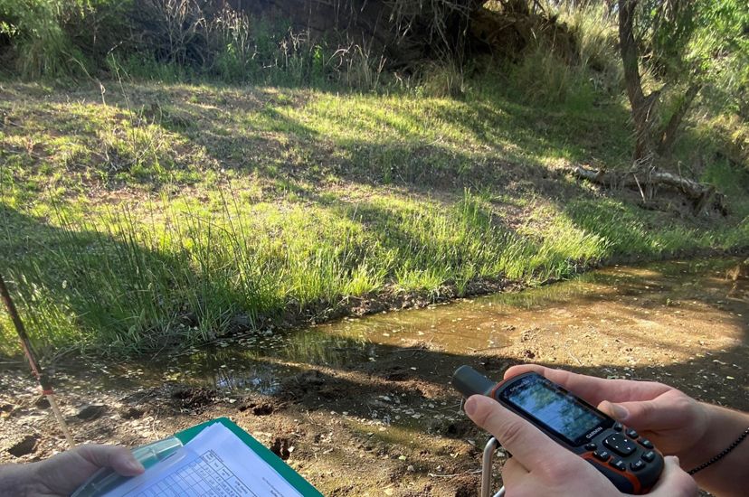 A person hand records data on a clipboard while another person reads information from a GPS unit.