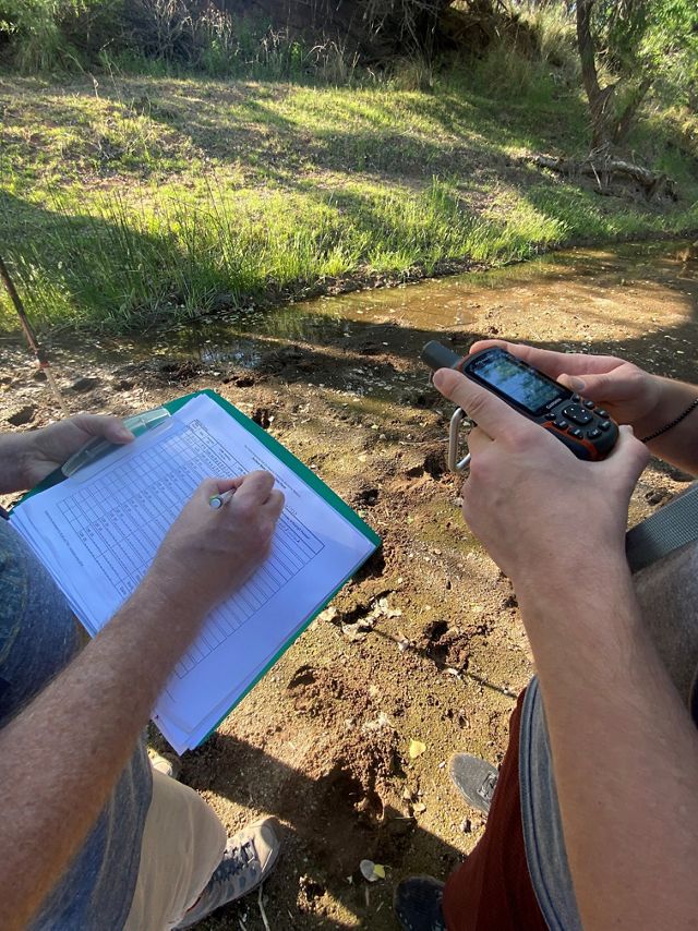 A person hand records data on a clipboard while another person reads information from a GPS unit.