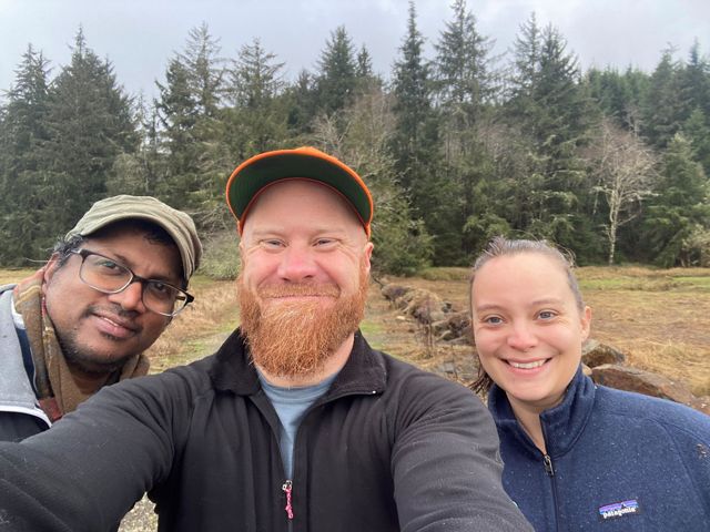 three people smiling at the camera with trees behind them.