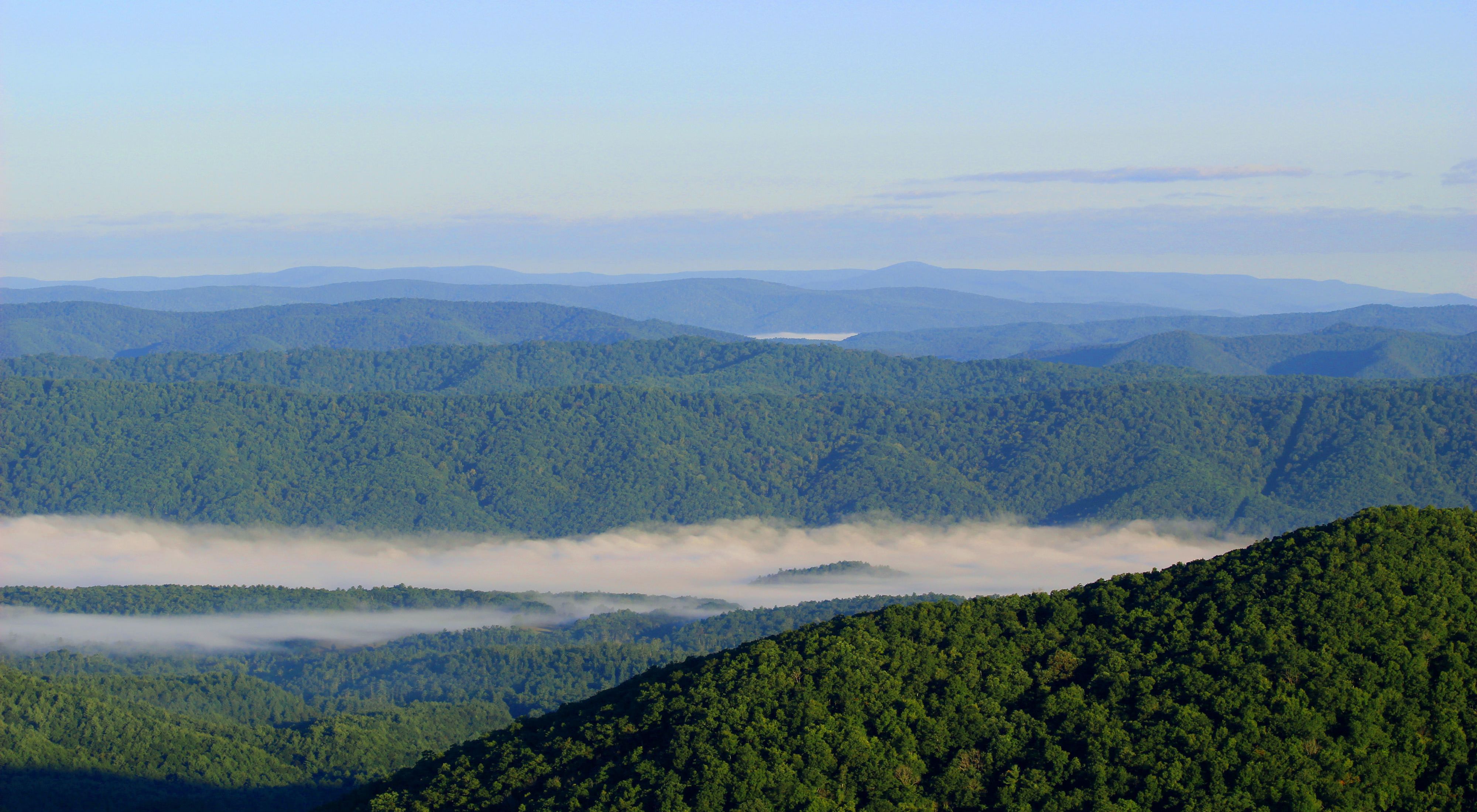 Aerial view of rolling green mountain ridge tops. A heavy bank of white fog shrouds the lower valley.