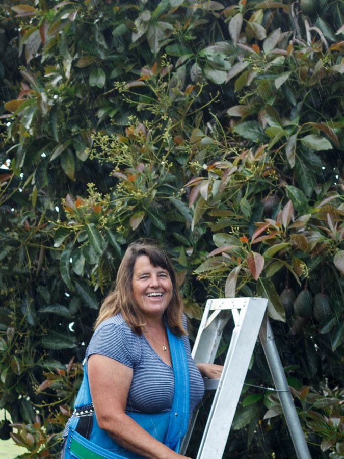 Person smiles at camera in front of avocado tree. 