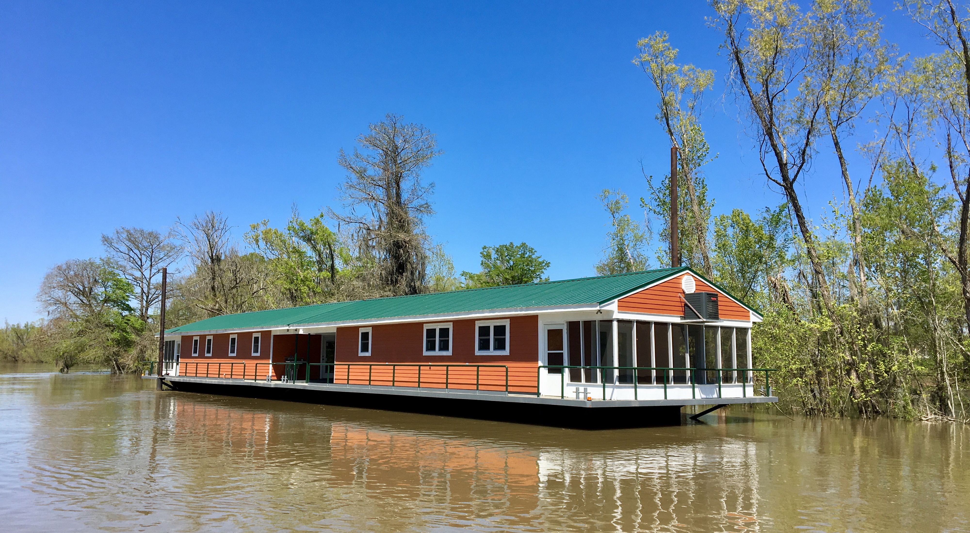 A large barge with a house-like structure on it floats on a flat, calm body of water with trees lining its banks.