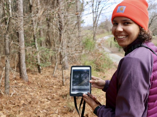Lauren Goodman stands by a forest tract holding a tablet.