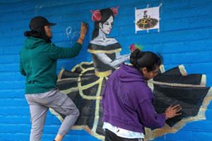 Two men paint a mural on a blue wall of a woman holding a bird.