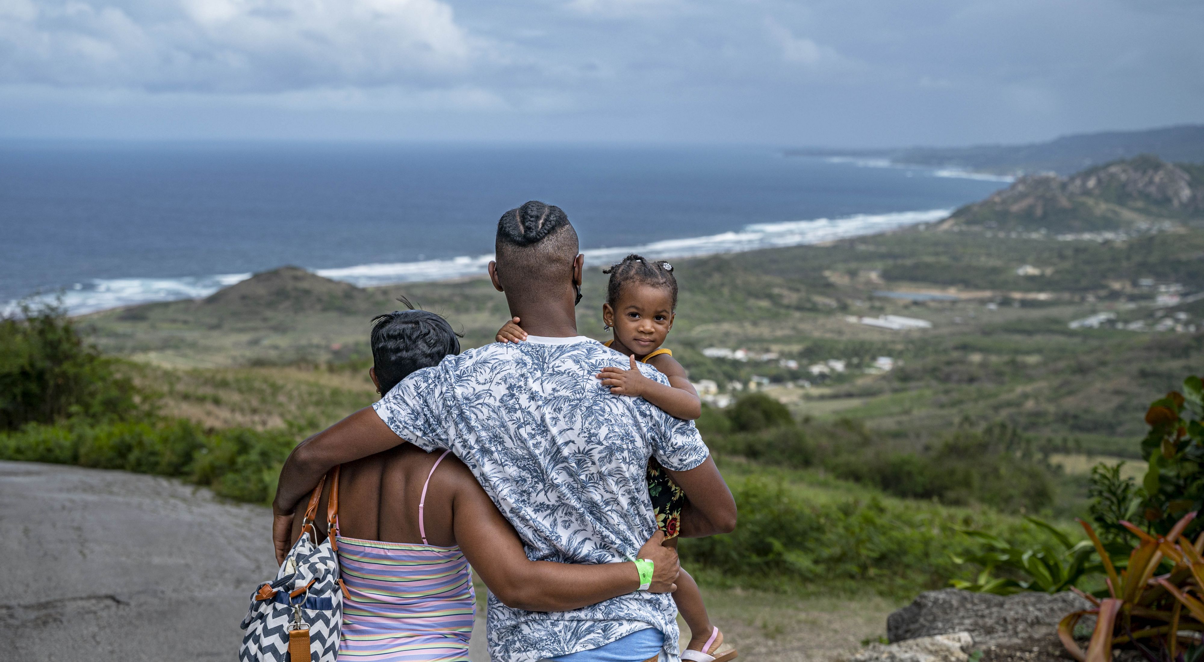 Fotografía de una familia (padre y madre con un niño en brazos) mirando hacia la cima de una colina de Barbados hacia el océano.