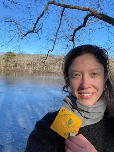 Bekah Myers stands outside in front of an icy pond surrounded by bare trees, holding a diamond-shaped yellow preserve boundary marker with the TNC logo.