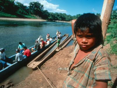 A child in the foreground looks directly in the camera as, in the background, a group of people board a large canoe on a river. 