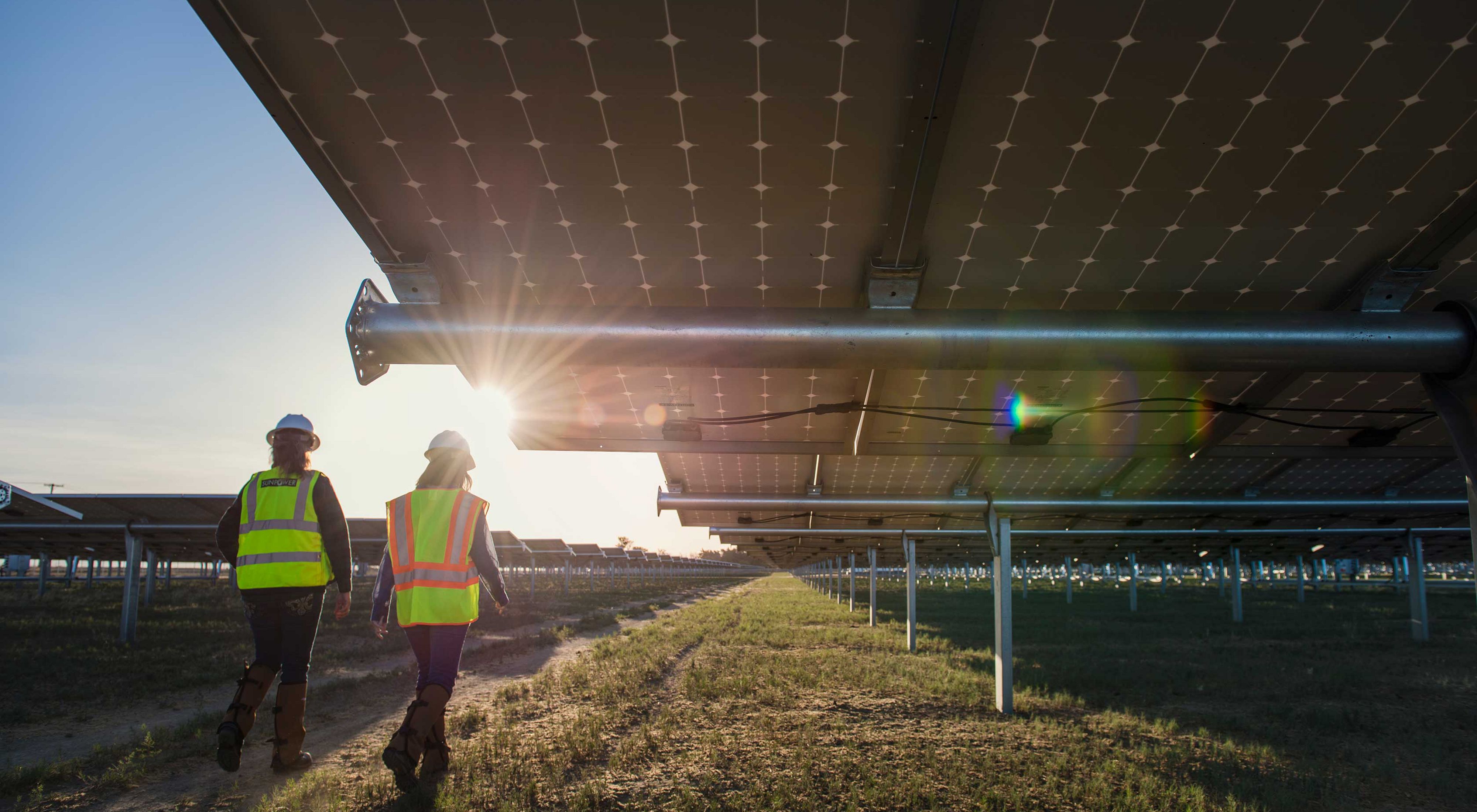 Two workers in hard hats and yellow vests walk underneath solar panels at a massive solar installation in California.