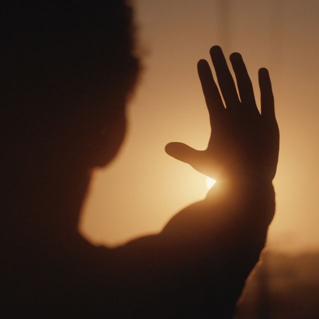 A silhouetted person holds their hand up to block the sun while the sky glows orange.